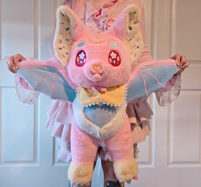 May include: A plush pink bat with large ears, star-shaped eyes, and light blue wings. The bat has a yellow bow tie and a heart-shaped nose. The wings are light blue with pink accents. The ears are cream-colored with colorful sprinkles.