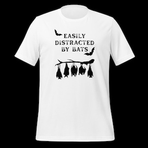 May include: A white short-sleeved t-shirt featuring a black graphic print. The design includes the text EASILY DISTRACTED BY BATS in a distressed font, accompanied by two flying bats and five bats hanging upside down from a tree branch.
