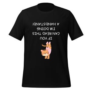 May include: A black short-sleeved t-shirt featuring an upside-down graphic of a tiger performing a handstand. Above the tiger, the text reads IF YOU CAN READ THIS I'M DOING A HANDSTAND! in white capital letters, also oriented upside down.