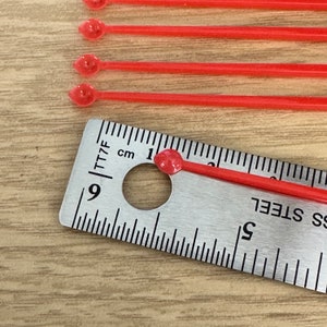 Pode incluir: Seis aplicadores de plástico vermelho com ponta arredondada, medidos com uma régua com marcas em polegadas e centímetros. A régua é marcada com o número 9 e o texto "TT7F".