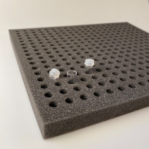 Op de afbeelding: Een zwarte schuimrubberen opberglade met meerdere ronde gaten, ontworpen om kleine flesjes of buizen te bewaren. Drie doorzichtige plastic flesjes zijn te zien in de lade, twee met doppen en één zonder.
