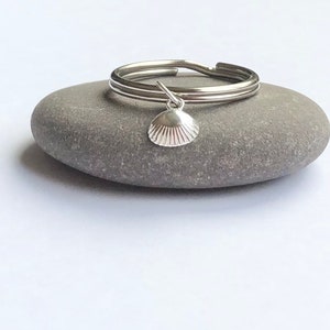 Op de afbeelding: Een zilveren ring met een kleine schelpbedel die eraan hangt. De ring ligt op een grijze steen.