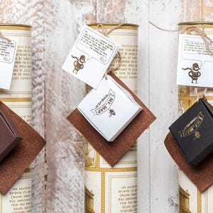 Peut inclure: Trois tubes cylindriques en carton avec des &eacute;tiquettes couleur cr&egrave;me comportant du texte et des bordures dor&eacute;es. Chaque tube poss&egrave;de une bo&icirc;te cadeau carr&eacute;e marron, blanche ou noire attach&eacute;e avec de la ficelle et une &eacute;tiquette blanche.