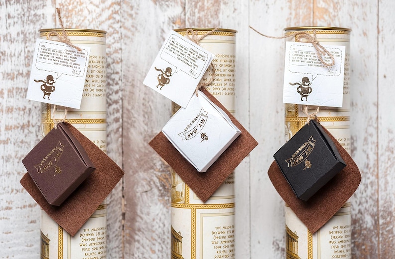 Peut inclure: Trois tubes cylindriques en carton avec des &eacute;tiquettes couleur cr&egrave;me comportant du texte et des bordures dor&eacute;es. Chaque tube poss&egrave;de une bo&icirc;te cadeau carr&eacute;e marron, blanche ou noire attach&eacute;e avec de la ficelle et une &eacute;tiquette blanche.