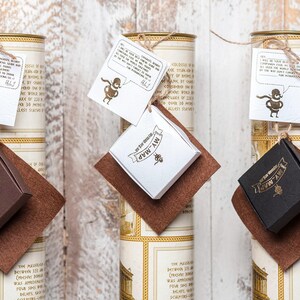 May include: Three cylindrical cardboard tubes with cream and gold vintage-style labels, each topped with a white square tag featuring a cartoon character and text, and a small square box in brown, white, or black attached to a brown felt square.