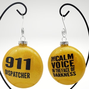 May include: Gold Christmas ornament with black text. One side reads "911 Dispatcher" and the other side reads "The Calm Voice in the Face of Darkness".