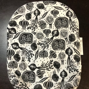 May include: Black and white oval fabric with a repeating pattern of vegetables. The vegetables include onions, radishes, and peas. The fabric has a white background with black outlines of the vegetables.