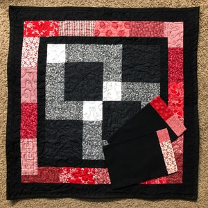 May include: A black and white quilt with a red and white border. The quilt is made of squares and has a geometric pattern. There are two black fabric squares with red and white borders on top of the quilt.