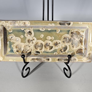 May include: A rectangular ceramic serving tray with a light brown glaze and a pattern of dark brown and white spots. The tray is on a black metal stand.