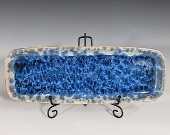 May include: A blue and white ceramic serving tray with a speckled glaze. The tray is rectangular and has a slightly rounded edge. It is displayed on a black metal stand.