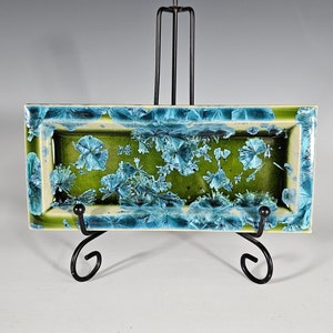 May include: A rectangular ceramic dish with a green glaze and blue crystalline patterns. The dish is on a black metal stand.