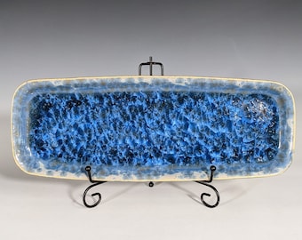 May include: A rectangular ceramic serving dish with a textured blue interior and a light blue and cream-colored border. The dish is displayed on a black metal stand, showcasing its unique design and craftsmanship.