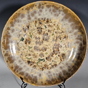 May include: A large, round ceramic plate with a unique, textured glaze. The plate features a central design of clustered, brown and beige patterns, surrounded by a lighter, speckled border. The rim is a warm, golden brown. The plate is displayed on a black metal stand.