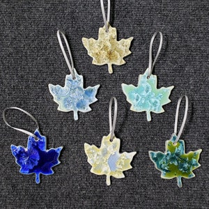 May include: Six ceramic maple leaf ornaments in various colors, including blue, green, and yellow, each with a unique crystalline pattern. The ornaments are strung with silver ribbons for hanging.