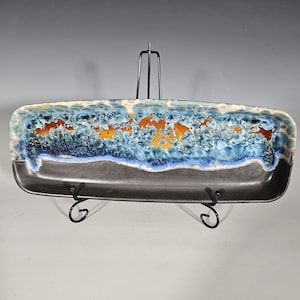 May include: Rectangular ceramic serving dish with a dark gray base and a top section featuring blue, orange, and white crystalline patterns. The dish is displayed on a black metal stand, showcasing its unique design.