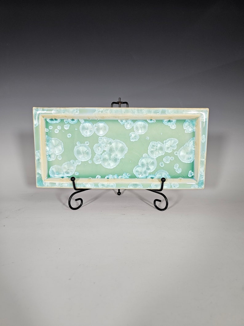 Puede incluir: Una bandeja rectangular de cer&aacute;mica con un esmalte verde claro y un patr&oacute;n moteado blanco. La bandeja est&aacute; sobre un soporte de metal negro.