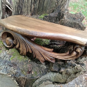 May include: A dark brown wooden wall shelf featuring a smooth, flat top surface and a decorative, hand-carved support bracket underneath. The bracket displays intricate, flowing scrollwork and leaf-like patterns carved from the same dark wood.