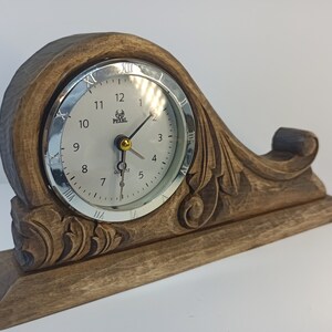 Puede incluir: Un reloj de madera con una esfera plateada y n&uacute;meros romanos negros. El reloj est&aacute; sobre una base de madera tallada con un dise&ntilde;o floral. La esfera del reloj dice "Pearl Quartz".