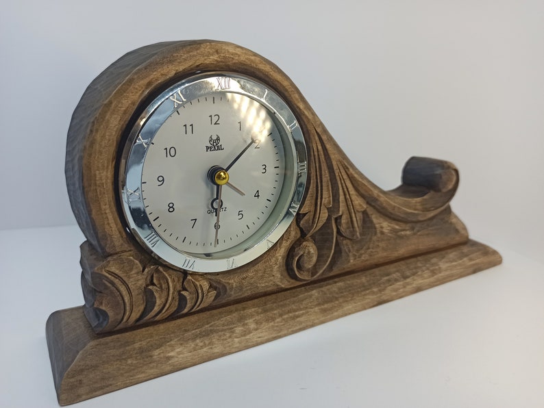Puede incluir: Un reloj de madera con una esfera plateada y n&uacute;meros romanos negros. El reloj est&aacute; sobre una base de madera tallada con un dise&ntilde;o floral. La esfera del reloj dice "Pearl Quartz".
