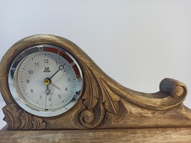 Puede incluir: Un reloj de madera con un dise&ntilde;o tallado y una esfera plateada. La esfera del reloj tiene agujas negras y n&uacute;meros romanos. La esfera del reloj tambi&eacute;n tiene las palabras "Quartz" y "Pearl" impresas en ella.