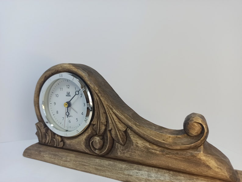 Puede incluir: Un reloj de madera con una base tallada y una esfera plateada. La esfera del reloj tiene n&uacute;meros romanos y un borde plateado. El reloj est&aacute; sobre un fondo blanco.
