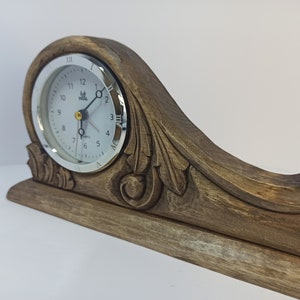 Puede incluir: Un reloj de madera con una esfera blanca y un borde plateado. El reloj est&aacute; montado sobre una base de madera tallada con detalles ornamentados. La esfera del reloj tiene n&uacute;meros romanos y un movimiento de cuarzo.