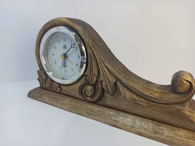 Puede incluir: Un reloj de madera con una esfera blanca y un borde plateado. El reloj est&aacute; montado sobre una base de madera tallada con detalles ornamentados. La esfera del reloj tiene n&uacute;meros romanos y un movimiento de cuarzo.