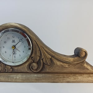 Puede incluir: Un reloj de madera con un dise&ntilde;o tallado y una esfera plateada. El reloj tiene n&uacute;meros romanos y una aguja de segundos dorada.