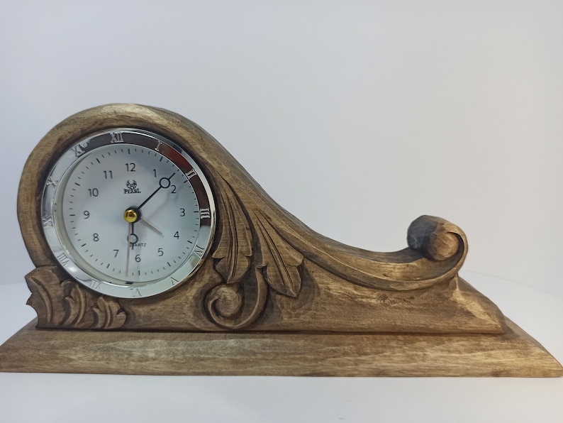 Puede incluir: Un reloj de madera con un dise&ntilde;o tallado y una esfera plateada. El reloj tiene n&uacute;meros romanos y una aguja de segundos dorada.