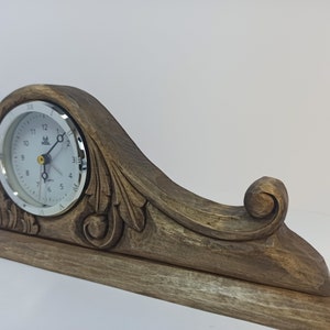 Puede incluir: Un reloj de madera con un dise&ntilde;o tallado y una esfera blanca. El reloj tiene un borde plateado y agujas negras.