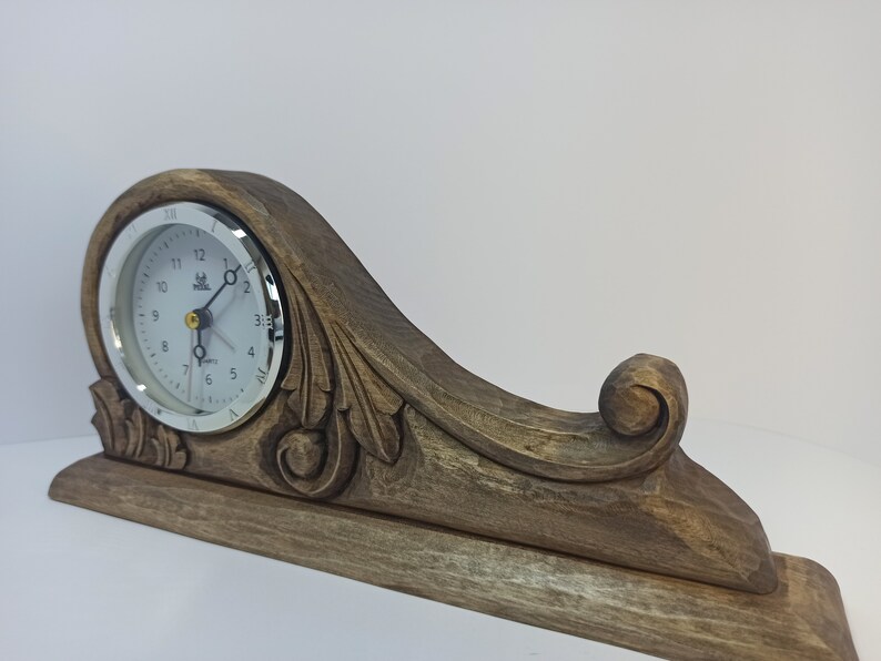 Puede incluir: Un reloj de madera con un dise&ntilde;o tallado y una esfera blanca. El reloj tiene un borde plateado y agujas negras.