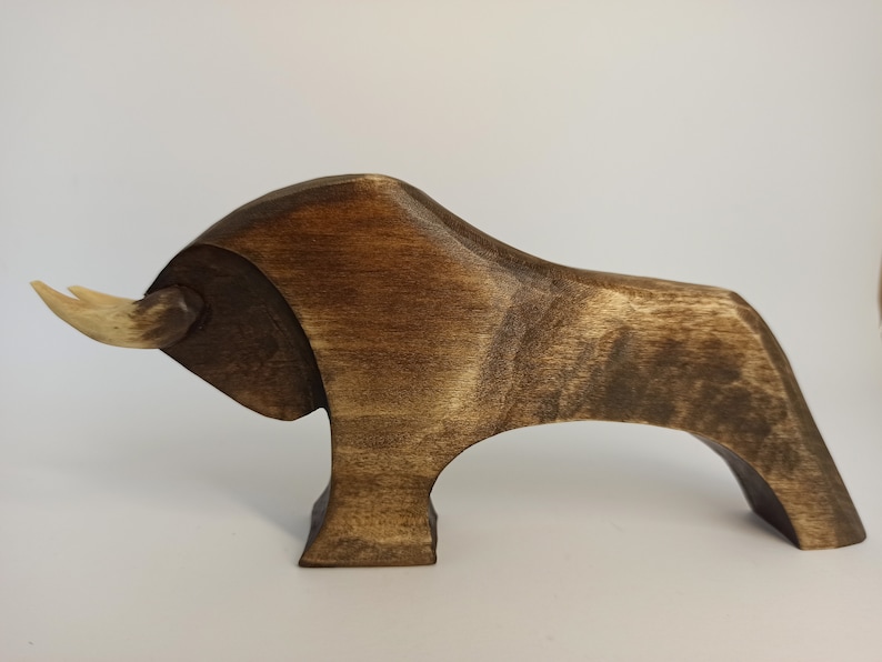 May include: A dark brown wooden sculpture of a bull with a light brown horn. The sculpture is stylized and has a simple design.