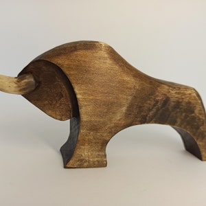 May include: A wooden sculpture of a bull with a light brown finish. The bull has a long, curved horn made of a lighter wood.