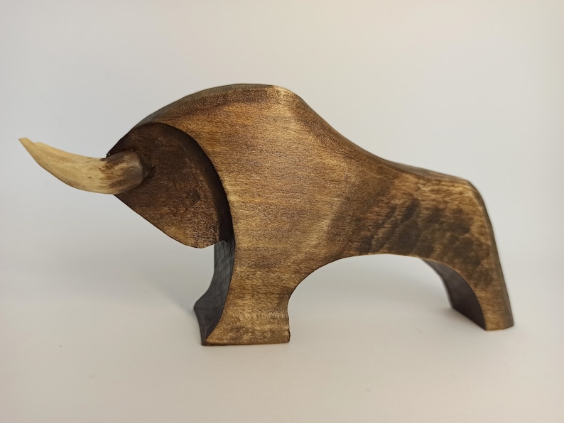 May include: A wooden sculpture of a bull with a light brown finish. The bull has a long, curved horn made of a lighter wood.