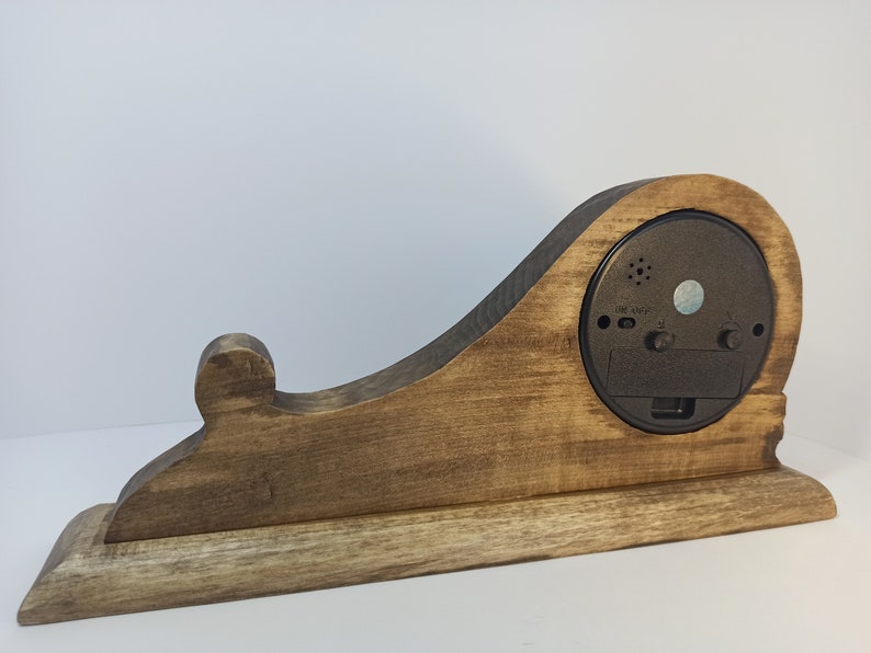 Puede incluir: Un reloj de madera con un acabado de tinte marr&oacute;n. El reloj tiene una forma &uacute;nica con una parte posterior curva y una base. La esfera del reloj no es visible en la imagen.