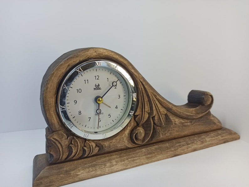 Puede incluir: Un reloj de madera con una base tallada y una esfera plateada. La esfera del reloj tiene n&uacute;meros romanos y una aguja de hora y minutos de color dorado. El reloj est&aacute; sobre un fondo blanco.