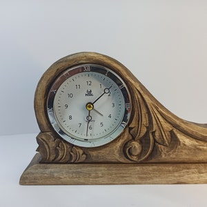 Puede incluir: Un reloj de madera con una esfera plateada y n&uacute;meros romanos. El reloj est&aacute; montado sobre una base de madera tallada con detalles intrincados.