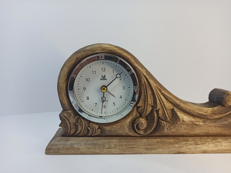 Puede incluir: Un reloj de madera con una esfera plateada y n&uacute;meros romanos. El reloj est&aacute; montado sobre una base de madera tallada con detalles intrincados.