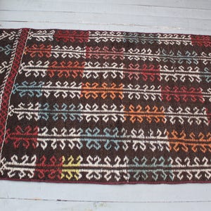 Op de afbeelding: Een bruin vloerkleed met een geometrisch patroon in tinten wit, rood, oranje, blauw en groen. Het vloerkleed heeft een franje rand.