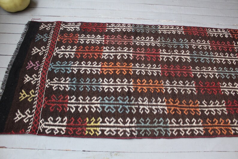 Op de afbeelding: Een bruin vloerkleed met een geometrisch patroon in tinten wit, rood, oranje, blauw en groen. Het vloerkleed heeft een franje rand.