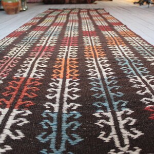 Op de afbeelding: Een lang, smal tapijt met een bruine achtergrond en een kleurrijk geometrisch patroon. Het patroon heeft witte, rode, oranje, blauwe en groene strepen met ingewikkelde ontwerpen.