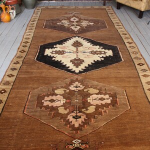 May include: A brown and beige rug with a geometric pattern. The rug has a central medallion with a black background and a white design. The rug has a border with a repeating pattern.