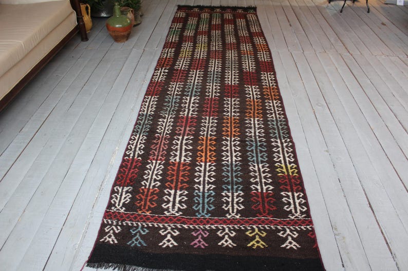 Op de afbeelding: Een lang, smal tapijt met een geometrisch patroon in tinten bruin, rood, wit en blauw. Het tapijt heeft aan beide uiteinden franjes.