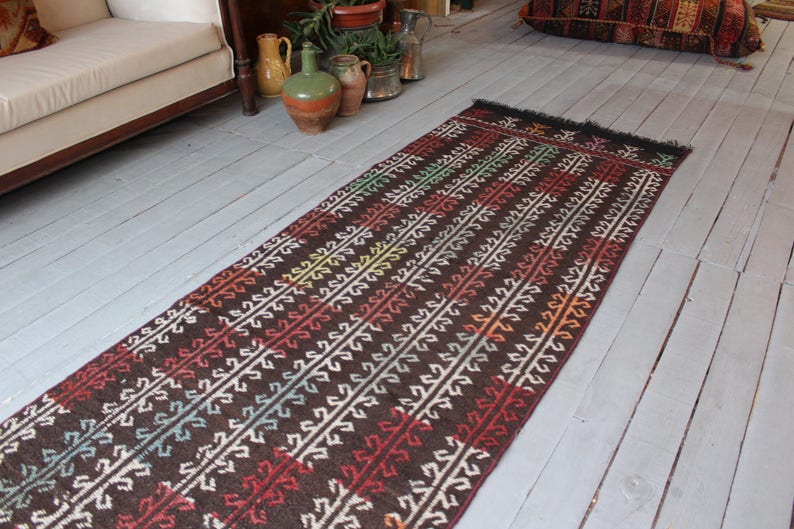 Op de afbeelding: Een lang, smal tapijt met een bruine achtergrond en een kleurrijk geometrisch patroon. Het tapijt heeft aan beide uiteinden franjes.
