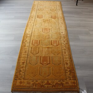 May include: A long, narrow, gold-colored rug with a geometric pattern. The rug has a brown border and a repeating pattern of diamond shapes.