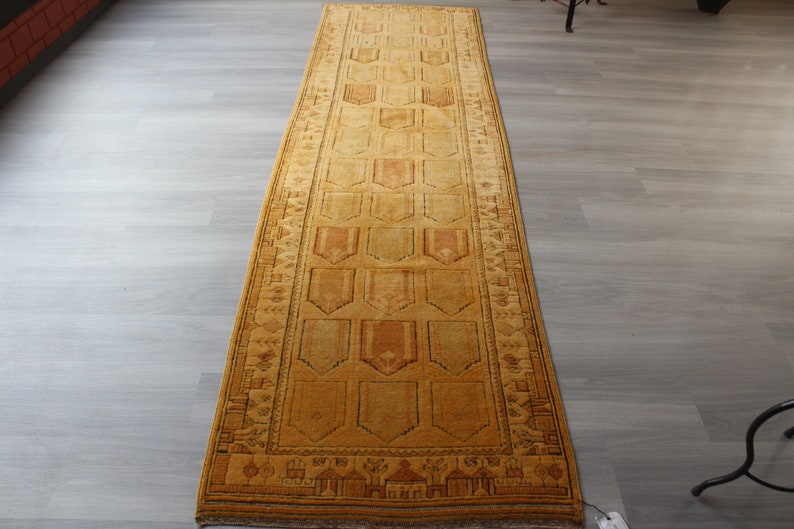 May include: A long, narrow, gold-colored rug with a geometric pattern. The rug has a brown border and a repeating pattern of diamond shapes.