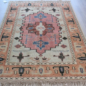Op de afbeelding: Een vintage tapijt met een crèmekleurige achtergrond en een roze en blauw bloemenontwerp. Het tapijt heeft een franje rand en ingewikkelde patronen.