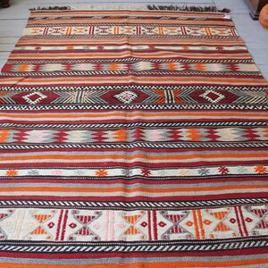 May include: A colorful striped rug with geometric patterns in shades of red, orange, white, black, and gray. The rug has a traditional woven design and is made of wool.