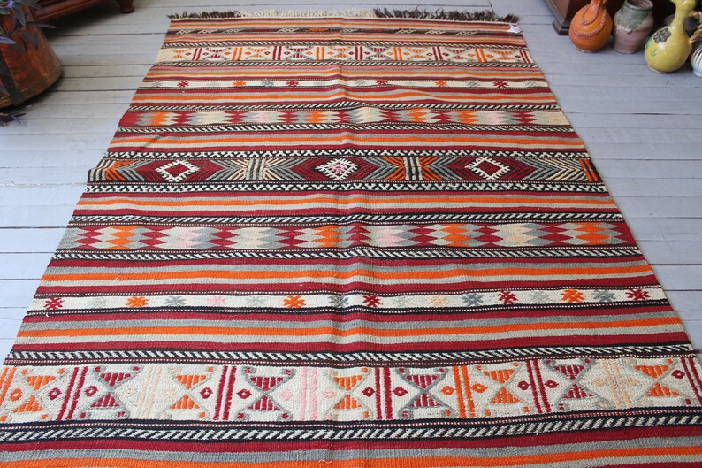 May include: A colorful striped rug with geometric patterns in shades of red, orange, white, black, and gray. The rug has a traditional woven design and is made of wool.