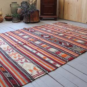 May include: A colorful striped rug with geometric patterns in shades of red, orange, white, and black. The rug has a fringe border and is laid out on a wooden floor.